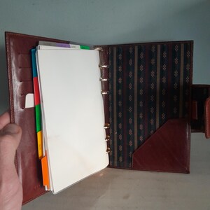 May include: Open brown leather binder with gold-colored rings, filled with white paper and colorful divider tabs. The interior features a patterned fabric lining and a pocket. The binder is open, revealing its contents.