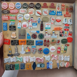 Op de afbeelding: Een verzameling vintage emaillepins met verschillende merken en logo's, waaronder Gala Koffie, Ivoorol, Medinos, Jant, Tempo en meer. De pins zijn in rijen op een bruin oppervlak gerangschikt.