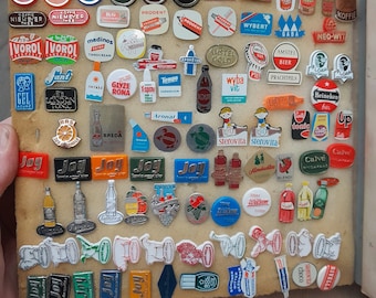 Op de afbeelding: Een verzameling vintage emaillepins met verschillende merken en logo's, waaronder Gala Koffie, Ivoorol, Medinos, Jant, Tempo en meer. De pins zijn in rijen op een bruin oppervlak gerangschikt.