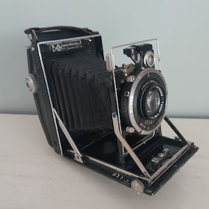 Può includere: Una macchina fotografica vintage nera e argentata con un soffietto e un obiettivo con la scritta "F. Schneider Kreuznach Tele-Xenar 1:4.5 105mm".