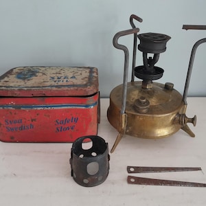 Könnte beinhalten: Ein antiker Svea Swedish Safety Stove mit einer roten und blauen Metallaufbewahrungsbox. Der Messingkocher hat einen schwarzen Brenner und Griff. Zusätzliche Teile sind ein kleiner Metallzylinder und zwei Metallwerkzeuge.