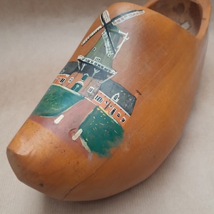 Op de afbeelding: Een houten klomp geschilderd met een scène van een Nederlandse windmolen en huizen. De klomp is bruin met groene, oranje en witte accenten.