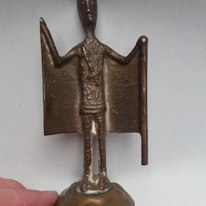 May include: A bronze-colored sculpture of a standing figure with outstretched arms and a rectangular shape on each side. The figure is on a round base. The sculpture has a textured surface and appears to be handmade.