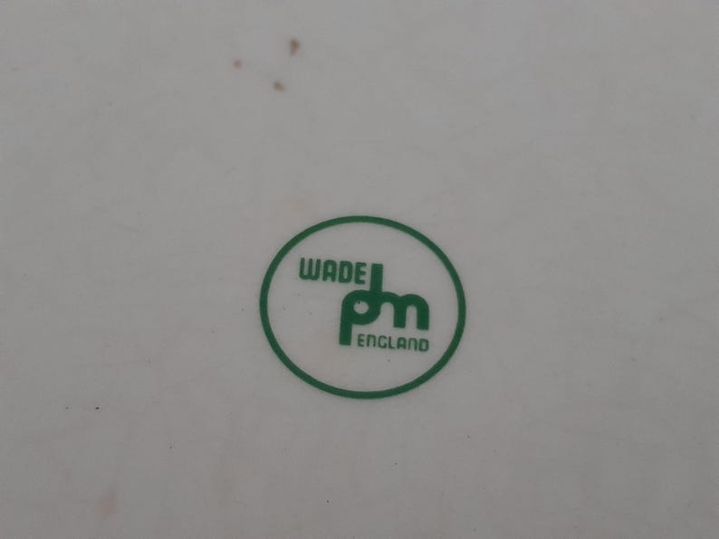 May include: A close-up of a white surface with a green circular logo. The logo features the words "WADE" and "pm" in a stylized font, with "ENGLAND" below. The logo is enclosed in a green circle.