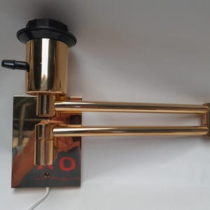 May include: A gold-toned, articulated wall-mounted lamp. The lamp features a rectangular base, a cylindrical body, and a double-arm design. The lamp has a black top and a black nozzle. The lamp is designed to be mounted on a wall.