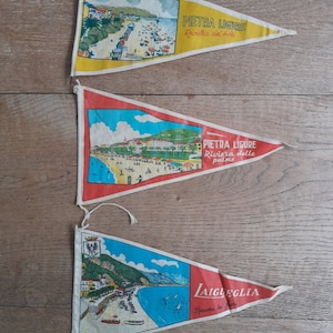 May include: Three vintage pennant flags featuring coastal scenes. The top flag is yellow with a beach scene and text "PIETRA LIGURE Riviera dei fiori". The middle flag is red with a beach scene and text "PIETRA LIGURE Riviera delle palme". The bottom flag is red with a coastal scene and text "LAIGUEGLIA Riviera dei fiori".