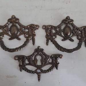 Könnte beinhalten: Drei antike Schubladengriffe aus Bronze. Zwei Griffe haben eine dekorative ovale Mitte und einen gebogenen Griff. Der dritte Griff hat ein Schlüsselloch-Design. Alle Griffe weisen aufwendige Details mit floralen und Blattakzenten auf.
