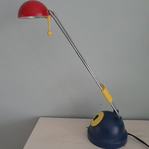 Può includere: Lampada da scrivania in stile retrò con paralume rosso, braccio cromato e base blu navy. La lampada presenta dettagli gialli sul braccio e sulla base e un piccolo cordino giallo. La lampada è collegata a una presa di corrente.