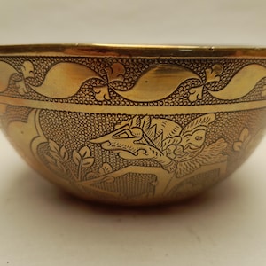 Puede incluir: Un cuenco dorado con intrincados diseños grabados. El cuenco presenta un patrón detallado de figuras estilizadas y motivos florales. El borde y la base del cuenco tienen un borde decorativo. El cuenco tiene un acabado liso y pulido.