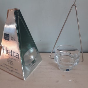 Puede incluir: Un portavelas de vidrio transparente con un marco colgante de metal plateado. El portavelas está etiquetado como "Hetta" y está hecho por Finn Crystal.