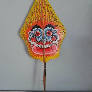 Puede incluir: Una marioneta de sombra de cuero pintada a mano con un rostro estilizado de ojos rojos grandes, boca ancha con dientes visibles y fondo amarillo con patrones de llamas en negro y naranja. Una varilla de madera está fijada verticalmente.