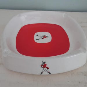 May include: White ceramic ashtray with a red center and a figure illustration. The ashtray has a square shape with rounded corners. The figure is wearing a red jacket and black pants.