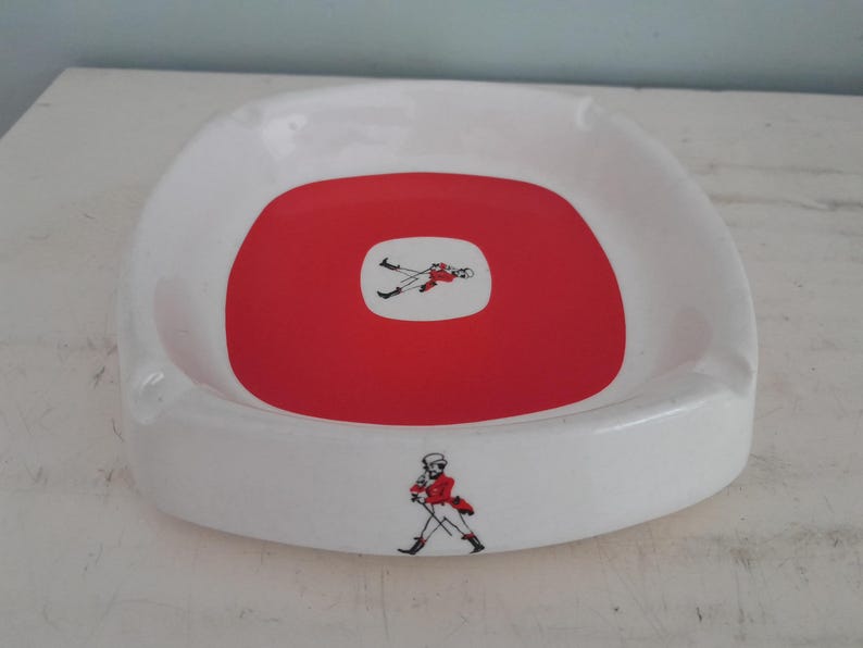 May include: White ceramic ashtray with a red center and a figure illustration. The ashtray has a square shape with rounded corners. The figure is wearing a red jacket and black pants.