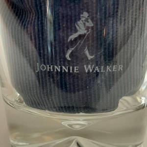 Pu&ograve; includere: Un bicchiere di vetro trasparente con un inserto in tessuto blu con il logo e il testo Johnnie Walker. Il logo raffigura un uomo con cappello e cappotto. Il bicchiere ha una base sfaccettata.
