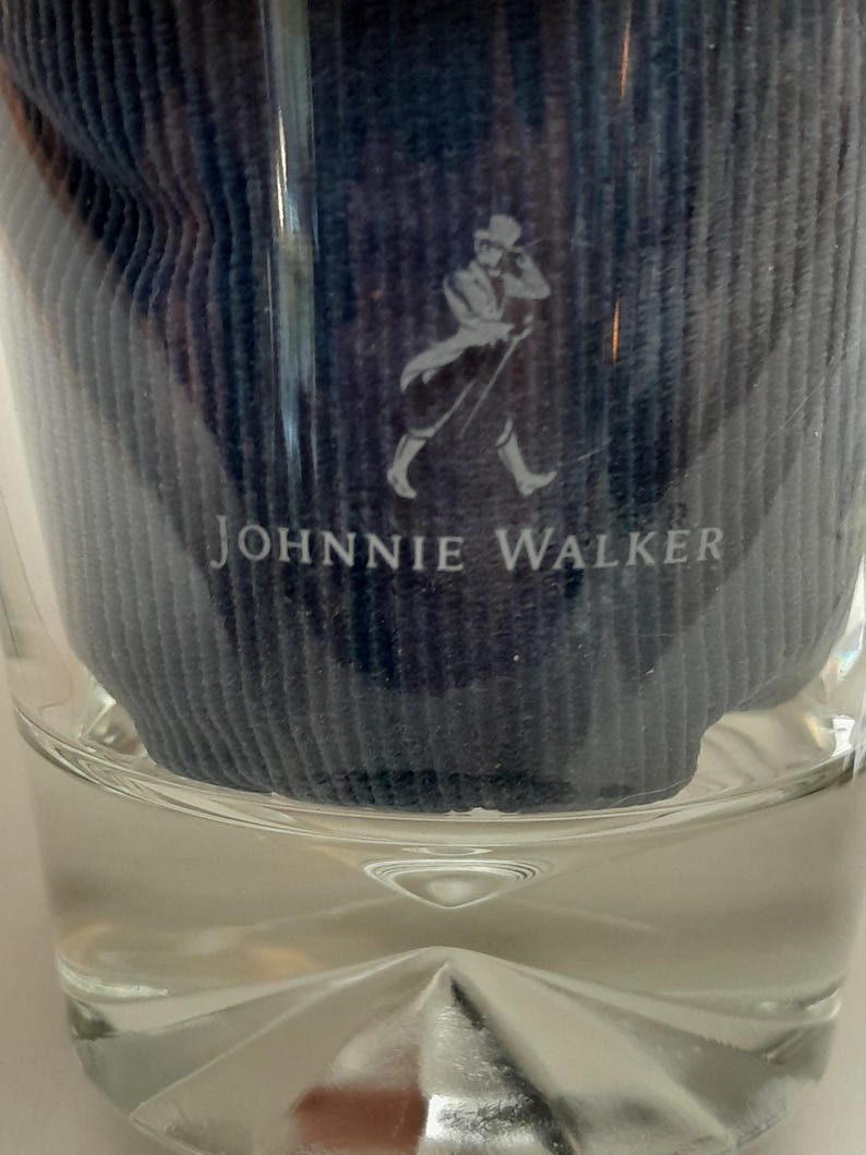 Pu&ograve; includere: Un bicchiere di vetro trasparente con un inserto in tessuto blu con il logo e il testo Johnnie Walker. Il logo raffigura un uomo con cappello e cappotto. Il bicchiere ha una base sfaccettata.
