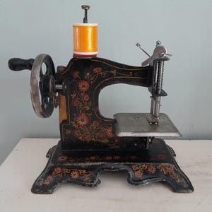 Op de afbeelding: Een vintage zwarte handnaaimachine met gedetailleerde bloemmotieven in rood, oranje en goud. De machine is voorzien van een zilverkleurig metalen handwiel met een zwarte hendel, een zilveren naaldstang en een felgele garenklos bovenop.