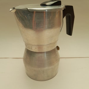 以下が含まれることがあります： 黒いプラスチック製のハンドルと蓋が付いた銀色のアルミニウム製のストーブトップエスプレッソメーカー。メーカーには側面に「Express」と「Italy」の文字が書かれています。