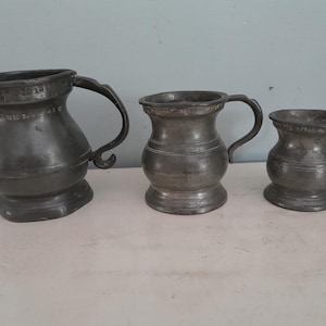 May include: Three antique pewter tankards of varying sizes. The tankards have a dark gray color and feature curved handles. The largest tankard has a bulbous base and a decorative rim. The middle tankard is smaller, and the smallest tankard is on the right.