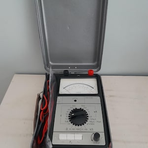 May include: A gray metal case containing a vintage electrical multimeter. The device has a black and gray face with dials, switches, and a needle gauge. Red and black wires are visible. The case is open, revealing the instrument.