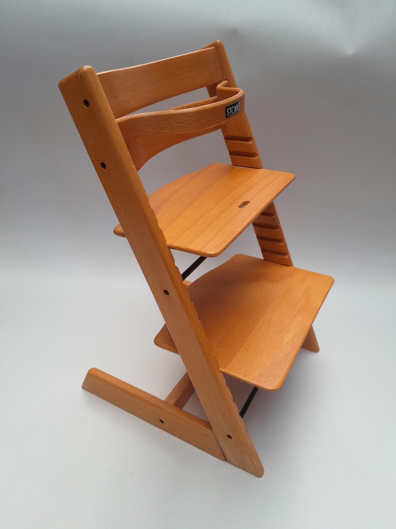 Puede incluir: Silla alta de madera de color marr&oacute;n claro con asiento y reposapi&eacute;s ajustables. La silla tiene un respaldo curvo y una barra de seguridad con la marca "Stokke". Dise&ntilde;ada para beb&eacute;s y ni&ntilde;os peque&ntilde;os, es ajustable.