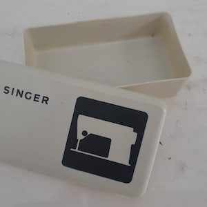 Op de afbeelding: Een witte plastic opbergdoos met het woord "SINGER" erop gedrukt. De doos heeft een zwart silhouet van een naaimachine aan de voorkant.