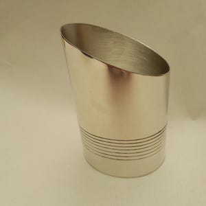 Puede incluir: Una taza de metal plateado con una parte superior inclinada y una serie de líneas horizontales cerca de la base. La taza tiene una superficie lisa y reflectante, y el interior también es plateado.