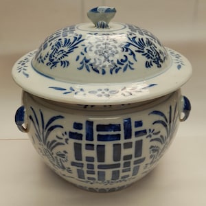 Puede incluir: Un tarro de cerámica blanco con un diseño floral azul y blanco. El tarro tiene dos asas y una tapa con un pomo en la parte superior. La tapa también tiene un diseño floral azul y blanco.