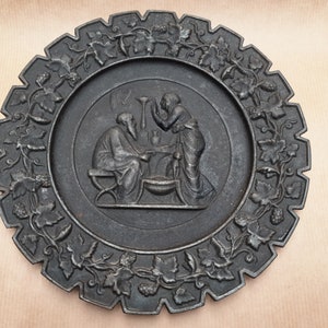 Op de afbeelding: Een zwarte gietijzeren wandplaat met een reliëf van twee figuren die aan een tafel zitten met een rankenrand.