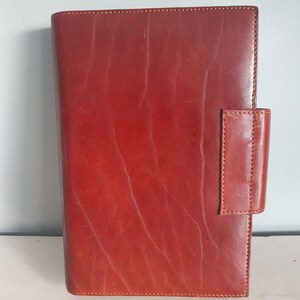May include: A reddish-brown leather journal with a rectangular shape. The journal has a stitched border and a flap closure on the right side. The leather has a slightly distressed appearance.
