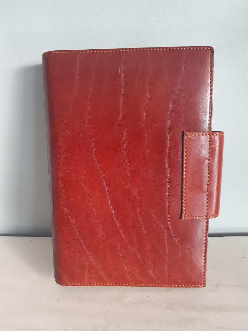 May include: A reddish-brown leather journal with a rectangular shape. The journal has a stitched border and a flap closure on the right side. The leather has a slightly distressed appearance.