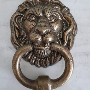 May include: A bronze-colored lion head door knocker. The lion's face is detailed with a mane, eyes, nose, and mouth. A circular handle hangs from the lion's mouth. The door knocker is made of metal and has an aged appearance.