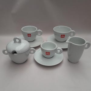 Può includere: Un set da espresso bianco con il logo "illy" in rosso. Il set comprende tre tazzine da espresso con piattini, una lattiera e una zuccheriera con coperchio e cucchiaio. Le tazze hanno piccole maniglie.