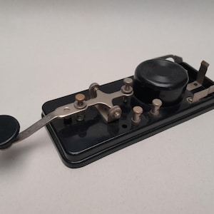 Op de afbeelding: Vintage zwarte telegraafsleutel met een ronde knop en een hendelarm. De sleutel heeft een rechthoekige basis met verschillende metalen componenten en een ronde zoemer. Een klassiek communicatie-apparaat.