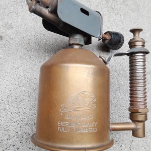 Könnte beinhalten: Eine vintage Lötlampe aus Messing mit zylindrischem Körper, grauem Metallbrennerkopf und geripptem Griff. Der Körper trägt die Gravur Neve, Holland, BLOWLAMP, EXCELLENT QUALITY, FULLY GUARANTEED sowie einen schwarzen Einstellknopf.