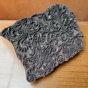 May include: A dark, weathered wooden printing block featuring an intricate, raised relief pattern of swirling floral vines, leaves, and small circular blossoms. The surface shows signs of age with worn edges and remnants of dark pigment in the crevices.