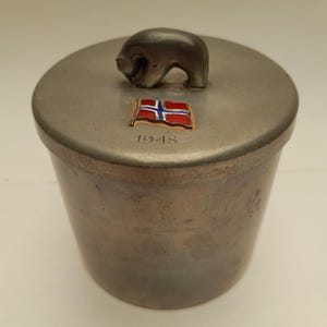Puede incluir: Un recipiente de metal plateado con una tapa que presenta una pequeña figura de oso y una bandera noruega con el año 1948 inscrito debajo.