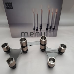 May include: A silver and black metal candle holder with six candle holders. The candle holder is designed in a modern style and is made by Menu.