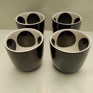 May include: A set of four rounded cups with a glossy black exterior and a white interior featuring large, irregular black circular spots. The cups have a smooth, handleless design and are arranged in a two-by-two grid formation.