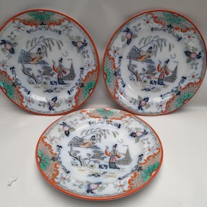 May include: Three round ceramic plates featuring a white base with a detailed transferware pattern. The design includes figures in traditional attire, willow trees, and architectural elements in orange, blue, and green, with a decorative orange rim.