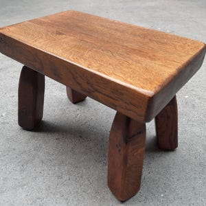 Op de afbeelding: Een kleine, handgemaakte houten kruk met een rechthoekige bovenkant en vier gebogen poten. De kruk is gemaakt van massief hout met een natuurlijke afwerking, waarbij de houtnerf zichtbaar is. De poten zijn stevig.