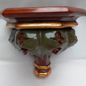 Pode incluir: Uma prateleira de parede com uma superfície superior de madeira lisa em castanho escuro. O suporte é decorado com uma secção central de padrões esculpidos em forma de folha verde-escura, acentuados por rebordos dourados metálicos.