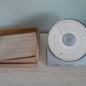 Może przedstawiać: Zabytkowe okrągłe suwmiarki z białą tarczą i szarym środkiem, zamontowane na srebrnym stojaku. Obok znajduje się otwarte kartonowe pudełko zawierające wydrukowany dokument. Suwak logarytmiczny to narzędzie do obliczeń matematycznych.