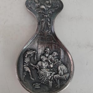 Puede incluir: Un reposacucharas plateado con un diseño en relieve detallado. El mango presenta un músico, mientras que el cuenco representa una reunión de personas. El reposacucharas es probablemente de metal y tiene una apariencia antigua.