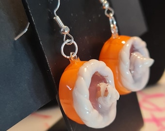 May include: A pair of drop earrings featuring miniature orange pumpkin-shaped charms topped with white twisted frosting and a central dollop of white cream, attached to silver-toned metal earring hooks.