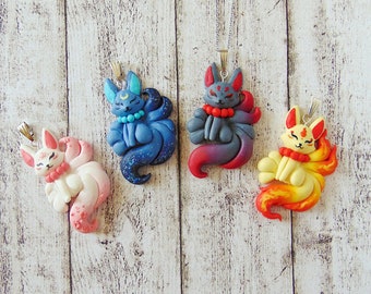 Op de afbeelding: Vier kleurrijke kitsune (vos) bedels met negen staarten, elk met een ander kleurenschema. De bedels zijn gemaakt van polymer clay en hebben een glanzende afwerking. De kitsune zijn afgebeeld in een zittende positie met hun staarten om hen heen gekruld.