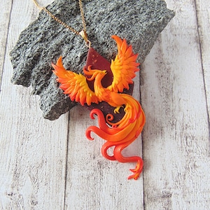 May include: A gold chain necklace with a phoenix pendant made of polymer clay. The phoenix is orange, yellow, and red with intricate details. The pendant is on a gray rock.