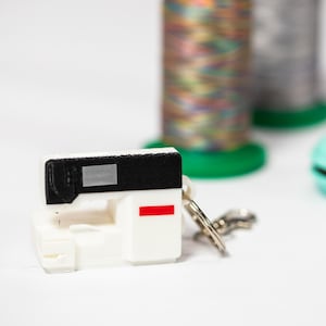 May include: A white miniature sewing machine keychain with a black top and a red stripe. Two spools of thread and a teal-colored item are in the background. The keychain has a silver clasp.