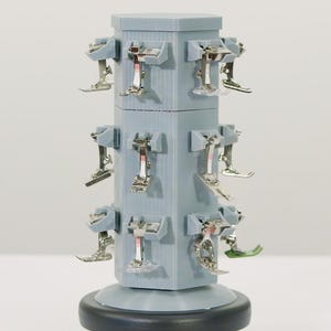 May include: A light blue, multi-tiered sewing machine foot display stand. The stand is cylindrical with a dark base and holds numerous silver sewing machine feet. The feet are arranged around the stand, showcasing various styles.
