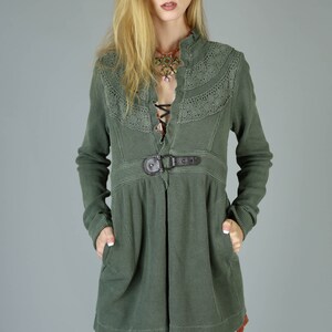 May include: A green waffle knit jacket with a lace trim and a brown belt. The jacket has a V-neck and long sleeves. The model is wearing the jacket with a pink velvet skirt and black tights.