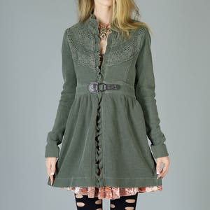 May include: A green waffle knit jacket with a lace trim, a brown belt, and a ruffled hem. The jacket is worn over a pink velvet skirt and black tights with cutouts.